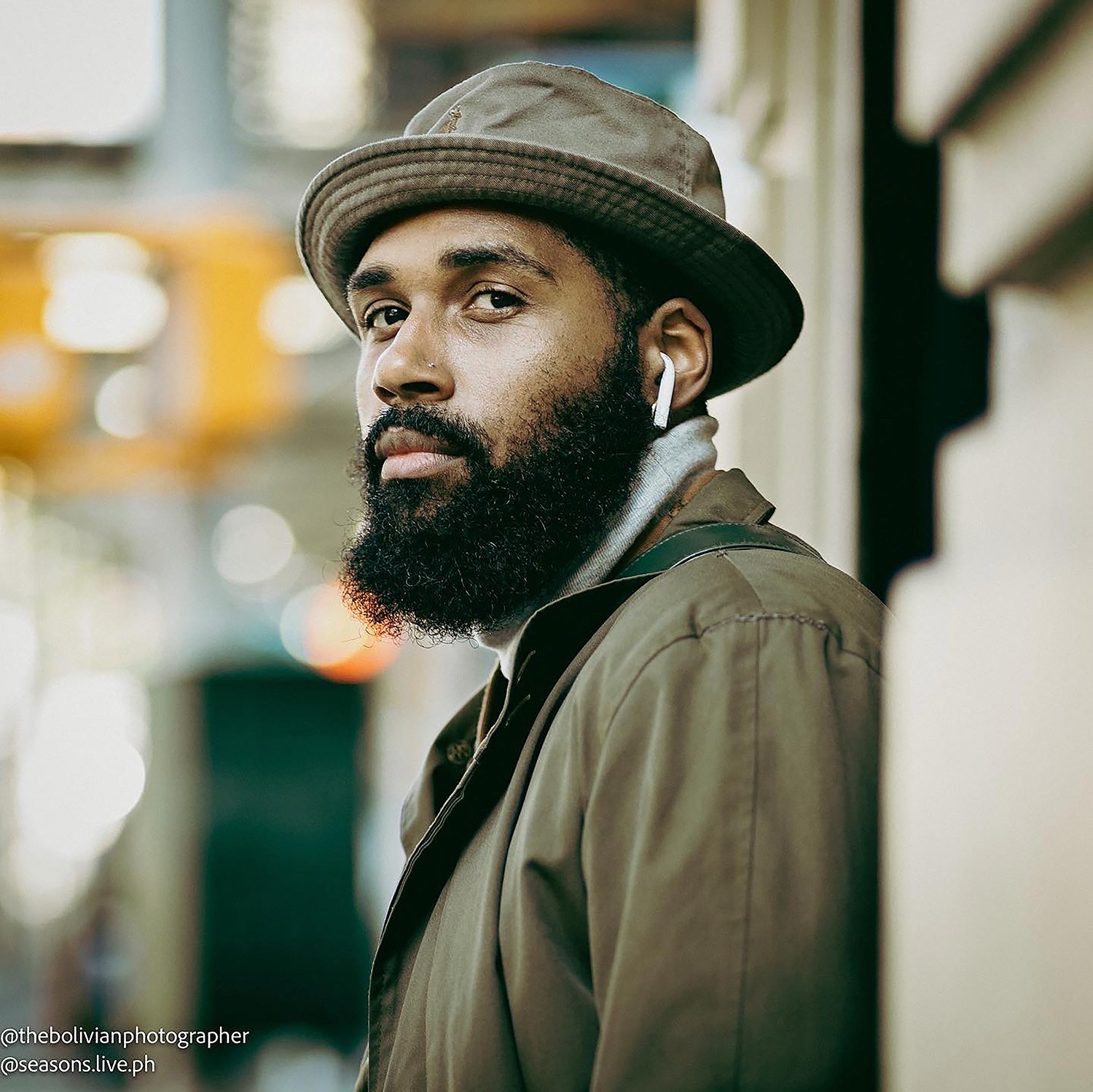 black man with beard, wearing hat and airpods, gazes slightly off camera. blurred cityscape behind him.